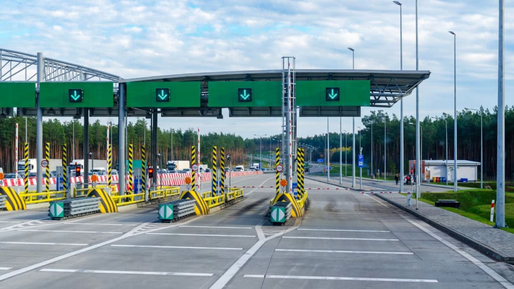 Cabines de pedágio com várias faixas abertas e sinalização verde, onde carros aguardam passagem em uma estrada cercada por árvores.