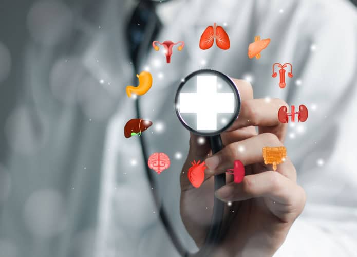 Uma pessoa com jaleco branco segura um estetoscópio com um símbolo de cruz brilhante no centro, cercado por ícones coloridos de órgãos humanos, representando cuidado médico completo.