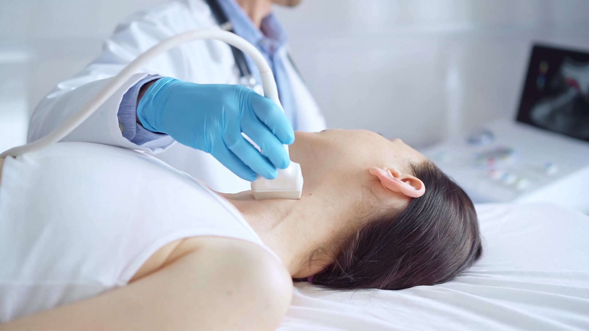 Uma profissional da saúde com luvas azuis realiza um exame de ultrassom no pescoço de uma paciente deitada. Ela desliza o aparelho sobre a região da tireoide enquanto, ao fundo, um monitor exibe imagens em tempo real do interior do pescoço.