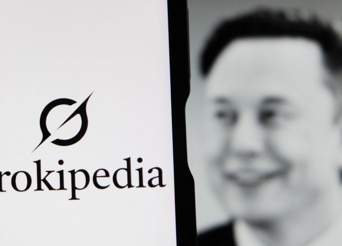 Celular com logomarca da Grokipedia na tela colocado na frente de monitor exibindo foto de Elon Musk