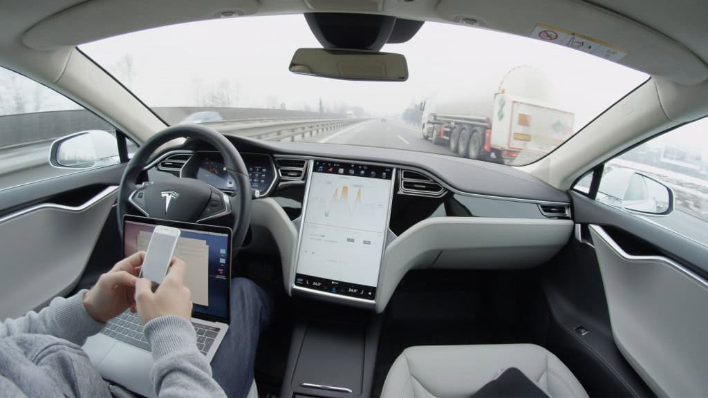 Pessoa com mexendo no celular e com notebook no colo sentada no banco do motorista de um carro da Tesla com Autopilot ativado e que está em movimento na rodovia