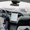 Pessoa com mexendo no celular e com notebook no colo sentada no banco do motorista de um carro da Tesla com Autopilot ativado e que está em movimento na rodovia