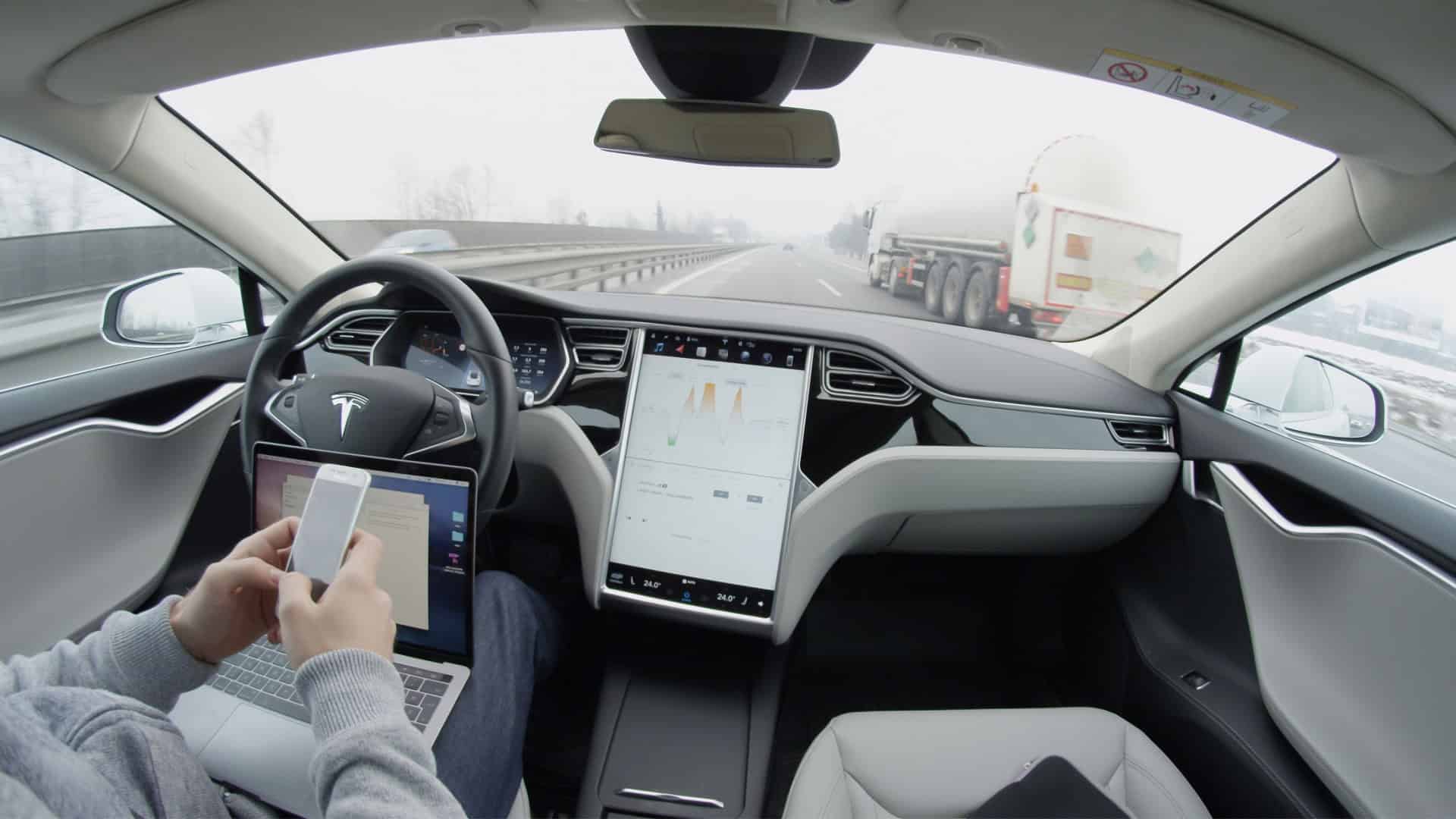 Pessoa com mexendo no celular e com notebook no colo sentada no banco do motorista de um carro da Tesla com Autopilot ativado e que está em movimento na rodovia