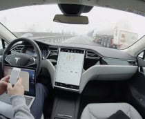 Pessoa com mexendo no celular e com notebook no colo sentada no banco do motorista de um carro da Tesla com Autopilot ativado e que está em movimento na rodovia
