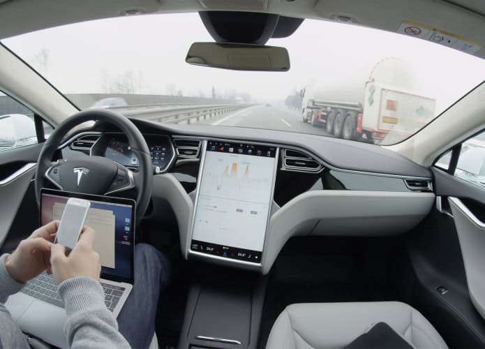 Pessoa com mexendo no celular e com notebook no colo sentada no banco do motorista de um carro da Tesla com Autopilot ativado e que está em movimento na rodovia