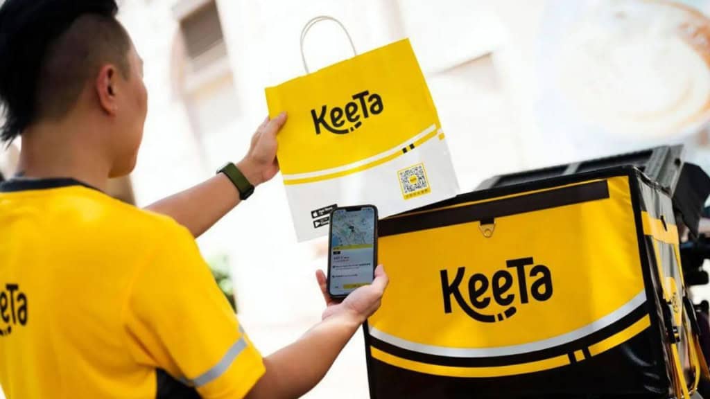 Entregador da Keeta segurando sacolinha da marca com uma mão e celular com aplicativo da marca aberto na outra, ao lado de moto com mochilona também da marca