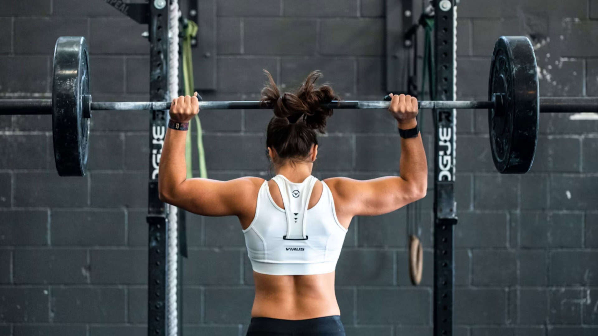 Mulher de costas, em frente a um apralho de academia, fazendo levantamento de peso.