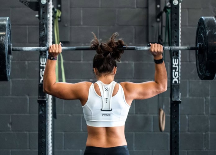 Mulher de costas, em frente a um apralho de academia, fazendo levantamento de peso.