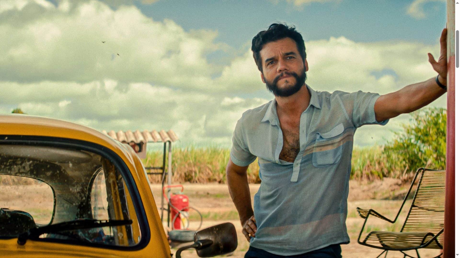 Wagner Moura encostado em uma parede ao lado de um carro