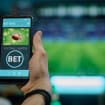 Ao fundo, borrado, uma partida de futebol passando em uma TV; à frente, a foto de uma bola de futebol americano e a palavra "bet" em um smartphone