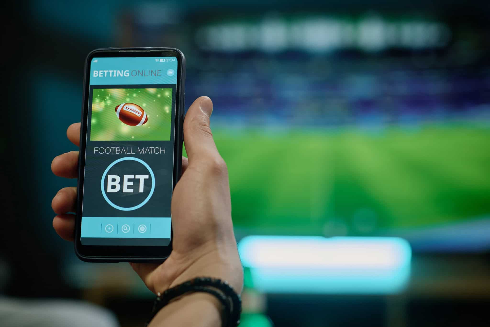 Ao fundo, borrado, uma partida de futebol passando em uma TV; à frente, a foto de uma bola de futebol americano e a palavra