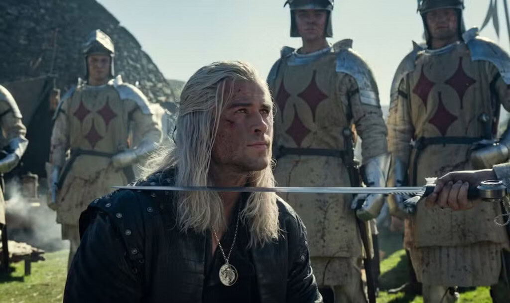 Cena de The Witcher mostra Geralt de Rívia ajoelhado, com uma espada encostada em seu ombro durante uma cerimônia de condecoração, cercado por soldados com armaduras medievais