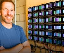 Homem que criou o sistema, sorrindo e de braços cruzados, com as baterias atrás, dispostas na parede, formando um quadrado