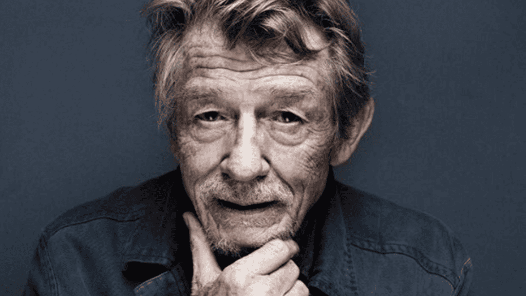 John Hurt om expressão serena e mão apoiada no queixo, olhando diretamente para a câmera, em referência ao tema “atores que mais morreram em filmes e séries”