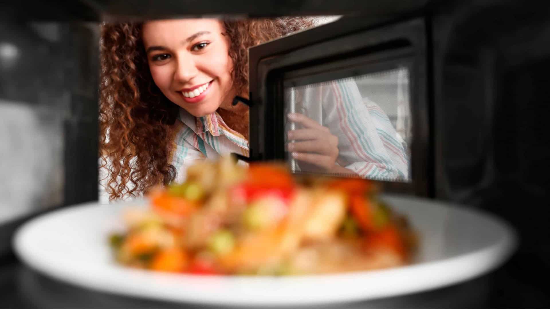 Visão de dentro de micro-ondas com prato de comida dentro e mulher olhando para ele e sorrindo