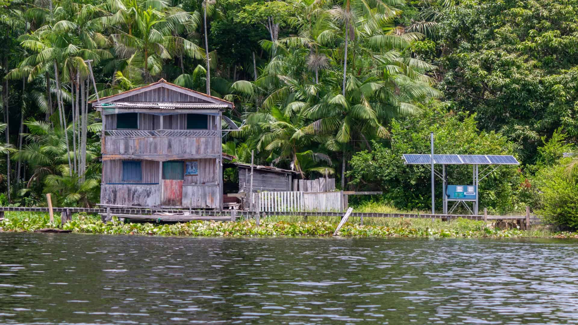 Casa de madeira em área ribeirinha da Amazônia ao lado de painéis solares entre vegetação densa, representando energia limpa e inclusão energética
