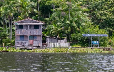 Casa de madeira em área ribeirinha da Amazônia ao lado de painéis solares entre vegetação densa, representando energia limpa e inclusão energética