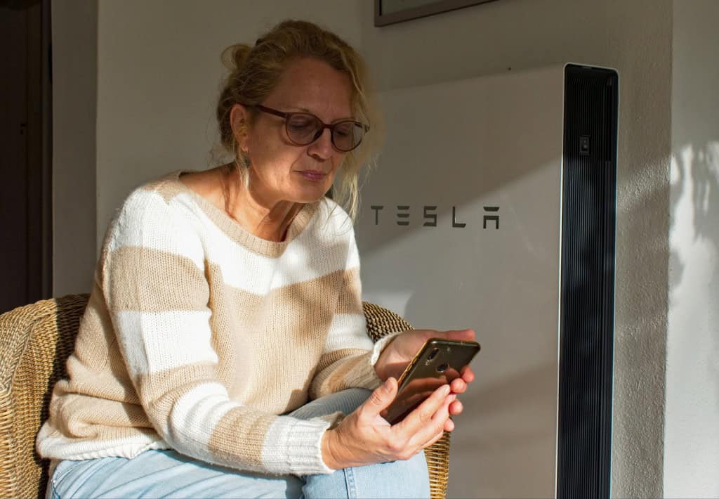 Ao fundo, uma caixa do TeslaPowerwall; à frente, uma mulher de óculos sentada, segurando um celular e olhando para ele