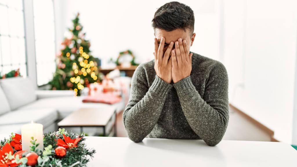 homem sentado com as mãos apoiando o rosto sobre a mesa, como se estivesse incomodado e ao fundo uma sala enfeitada para o natal 