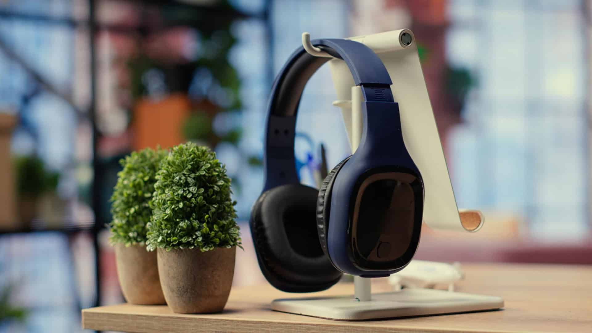 fone de ouvido preto apoiado em um suporte claro em cima de um móvel de madeira com duas plantinhas do lado