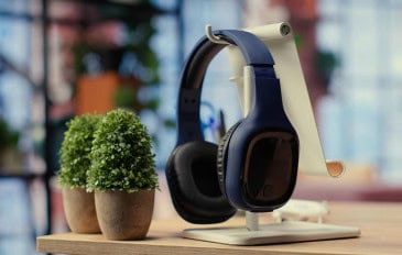 fone de ouvido preto apoiado em um suporte claro em cima de um móvel de madeira com duas plantinhas do lado