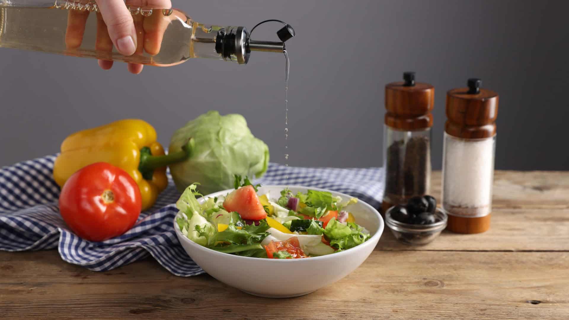 Uma mão derrama líquido de uma garrafa sobre uma salada colorida com folhas, tomates, pepinos e pimentões.