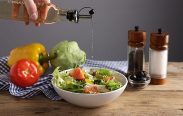 Uma mão derrama líquido de uma garrafa sobre uma salada colorida com folhas, tomates, pepinos e pimentões.