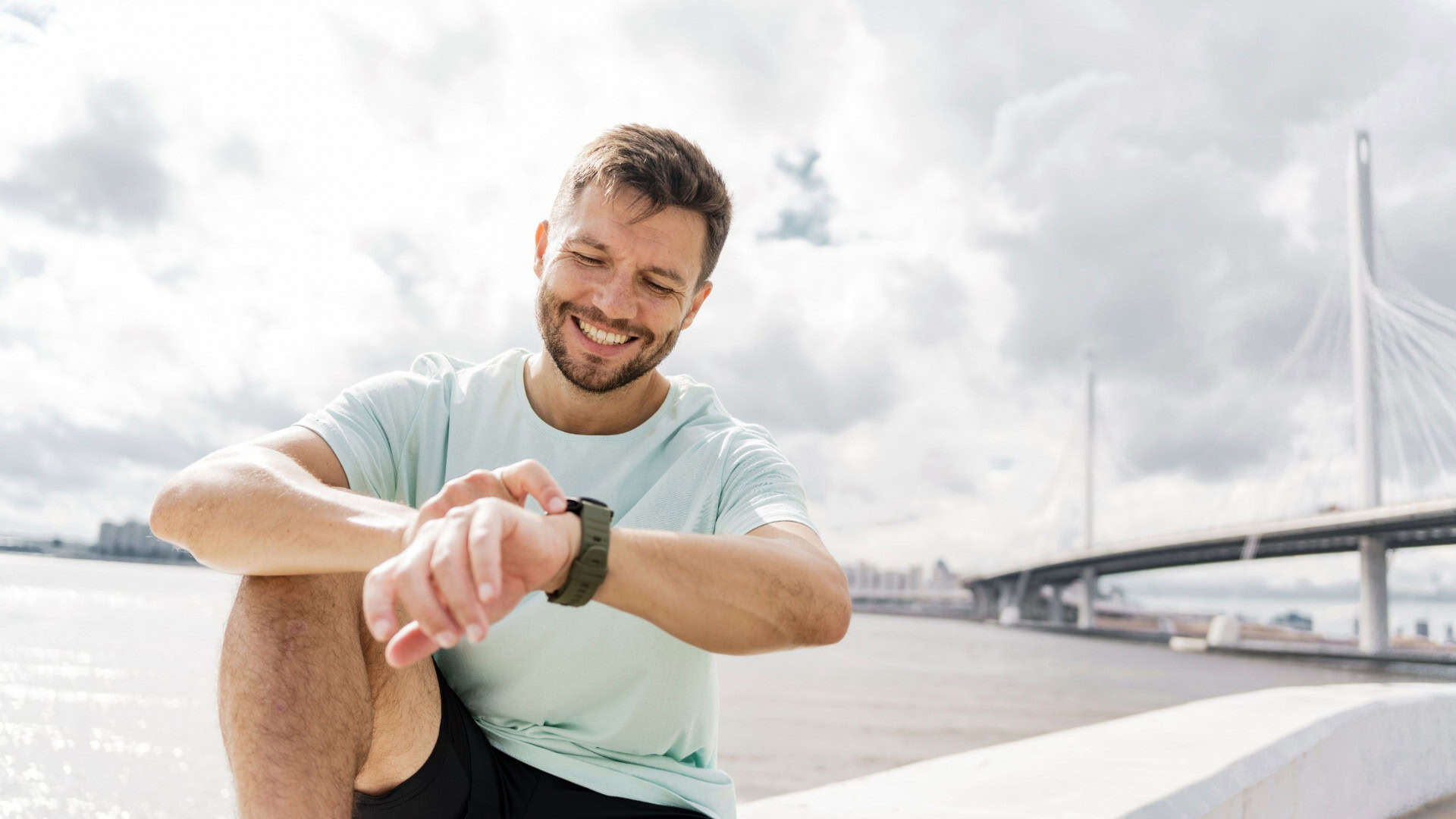 Pessoa sentada ao ar livre, sorrindo enquanto olha para o Apple Watch no pulso, com uma ponte moderna ao fundo e clima nublado.