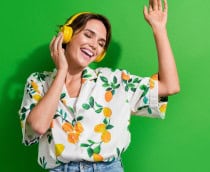 Mulher de cabelo curto e castanho, sorrindo com os olhos fechados, curte música com fones amarelos vibrantes. Veste camisa branca estampada com limões e folhas, calça jeans, e está diante de um fundo verde intenso. A pose transmite alegria e leveza.