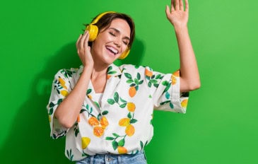 Mulher de cabelo curto e castanho, sorrindo com os olhos fechados, curte música com fones amarelos vibrantes. Veste camisa branca estampada com limões e folhas, calça jeans, e está diante de um fundo verde intenso. A pose transmite alegria e leveza.