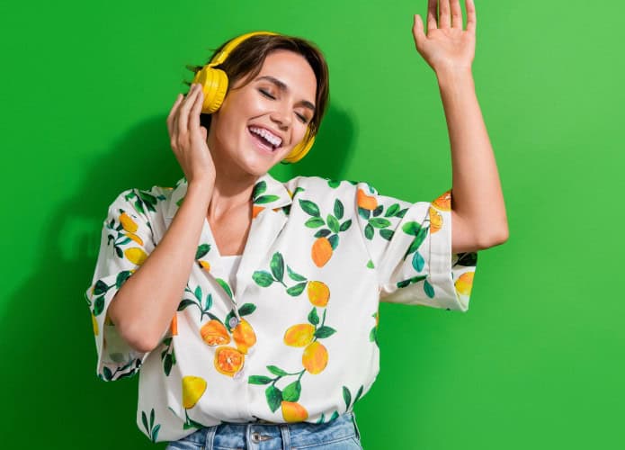 Mulher de cabelo curto e castanho, sorrindo com os olhos fechados, curte música com fones amarelos vibrantes. Veste camisa branca estampada com limões e folhas, calça jeans, e está diante de um fundo verde intenso. A pose transmite alegria e leveza.