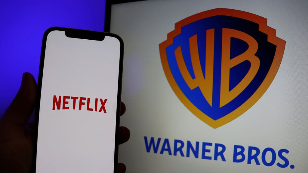 Pessoa segurando celular com logomarca da Netflix na tela; ao fundo, um monitor exibe logomarca da Warner Bros.