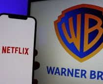 Pessoa segurando celular com logomarca da Netflix na tela; ao fundo, um monitor exibe logomarca da Warner Bros.