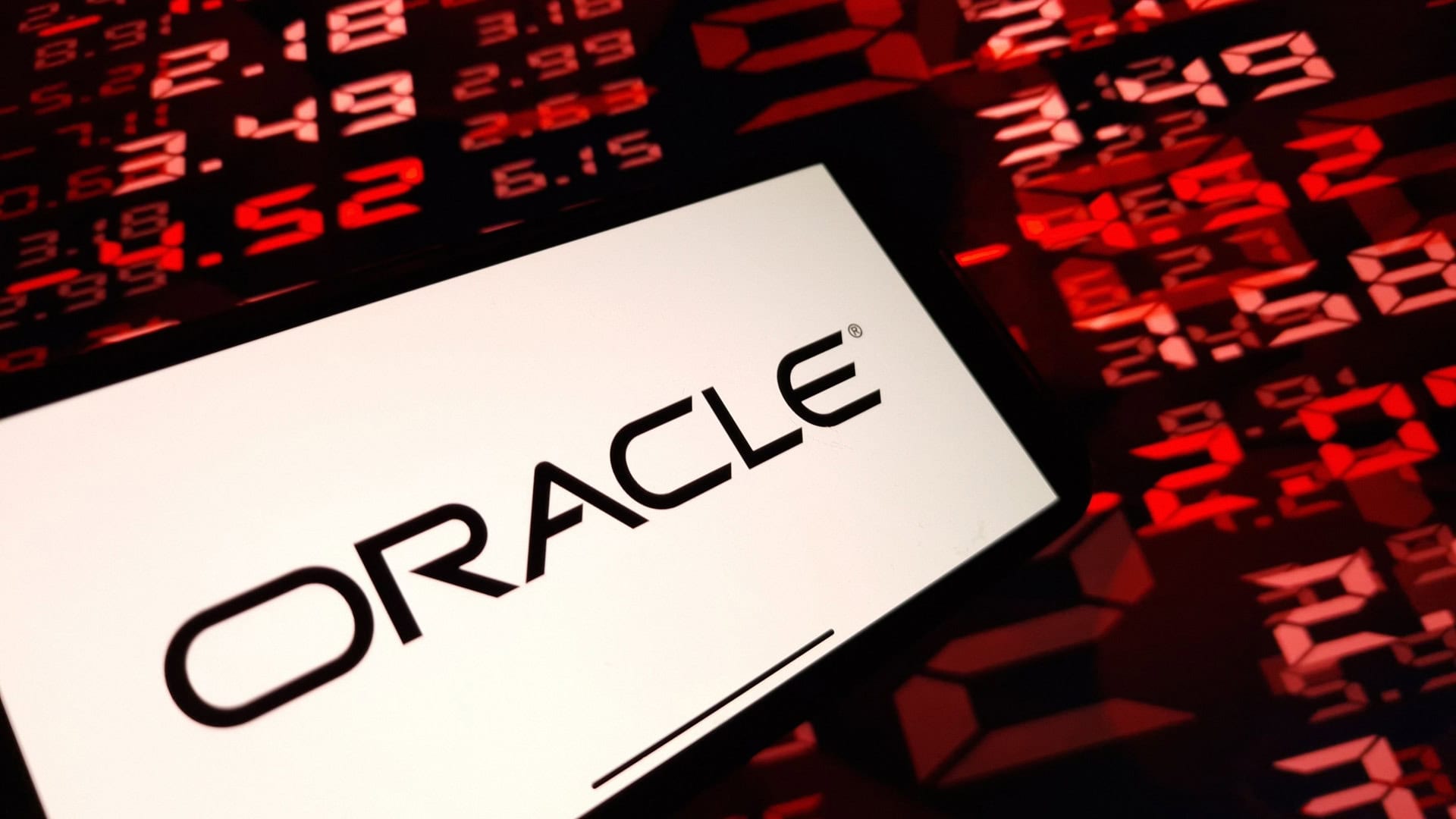 Pessoa segurando celular com logomarca da Oracle na tela na frente de tela mostrando números de ações financeiras vermelhos