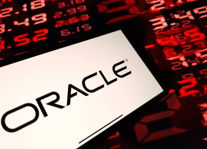 Pessoa segurando celular com logomarca da Oracle na tela na frente de tela mostrando números de ações financeiras vermelhos