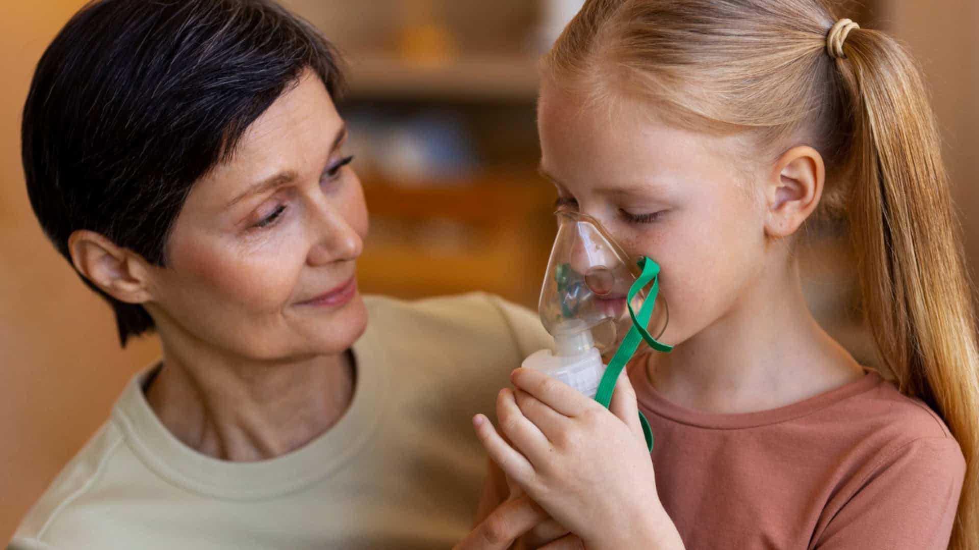 uma mãe fazendo nebulização em sua filha criança, dentro de casa, com os móveis desfocados ao fundo.