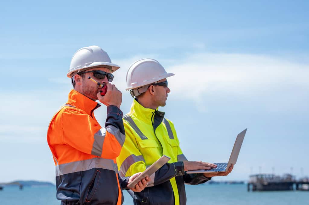 Dois homens com aparatos de segurança; o da esquerda está segurando um rádio comunicador próximo à boca; o da direita segura um notebook