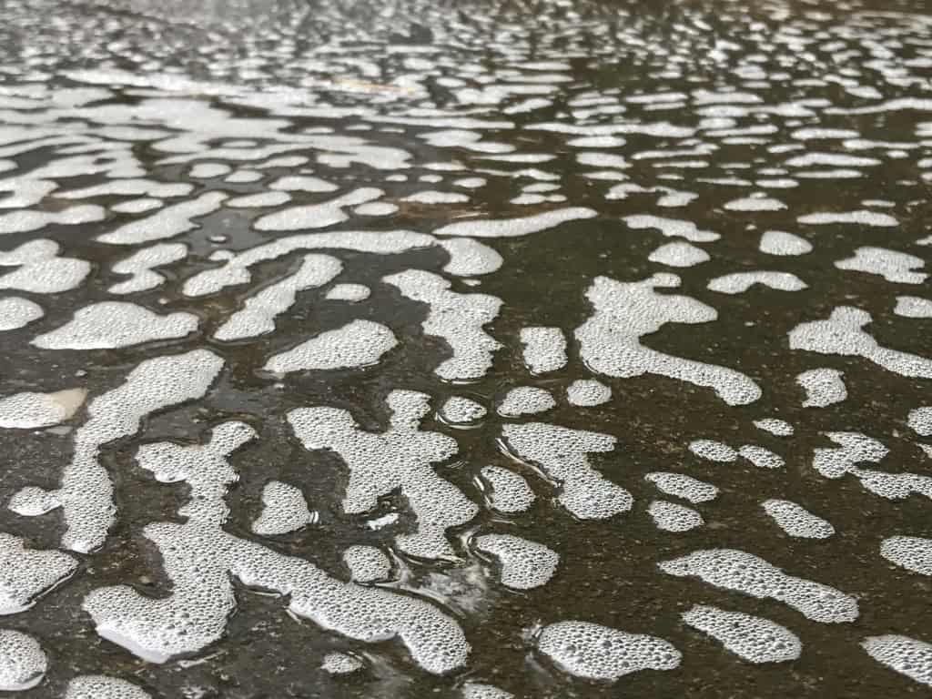 Superfície de água escura coberta por manchas de espuma branca, representando contaminação e poluição química em ambientes aquáticos