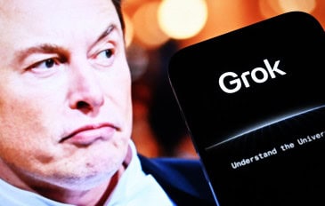 Celular mostrando logomarca do Grok; ao fundo, um monitor exibe uma foto de Elon Musk como se ele olhasse para o aparelho