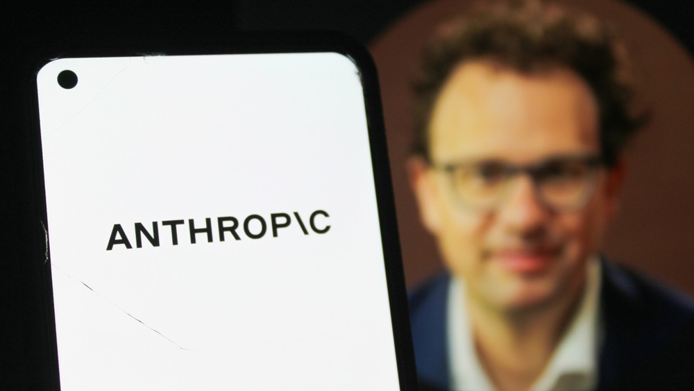 Logo da Anthropic em primeiro plano em um smartphone; ao fundo, imagem desfocada de Dario Amodei