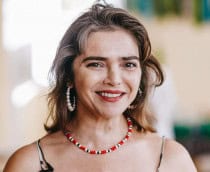 imagem mostra um close-up da atriz Titina Medeiros, sorrindo para a câmera em um fundo desfocado