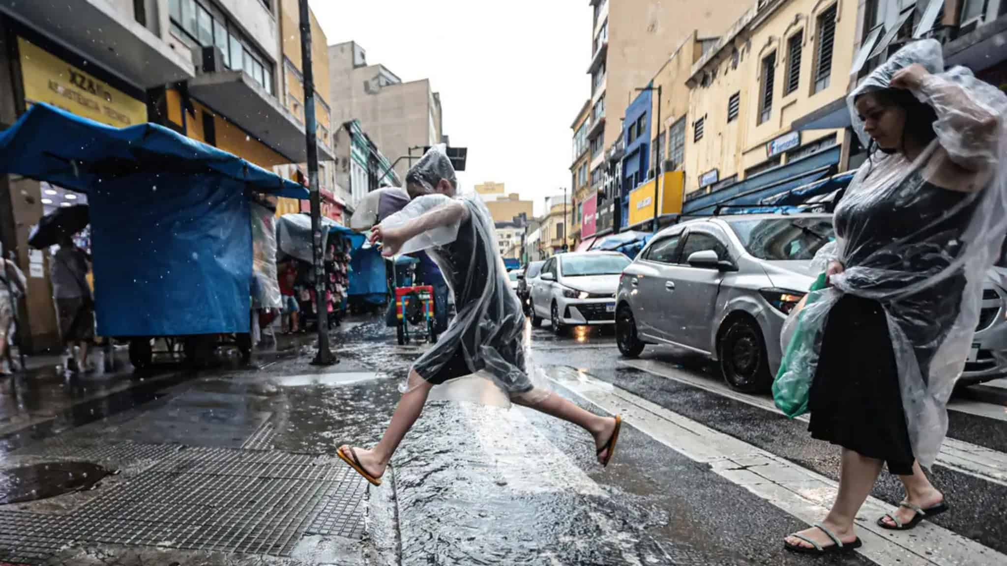 Mulher e criança vestindo capa de chuva e atravessando rua; criança está pulando
