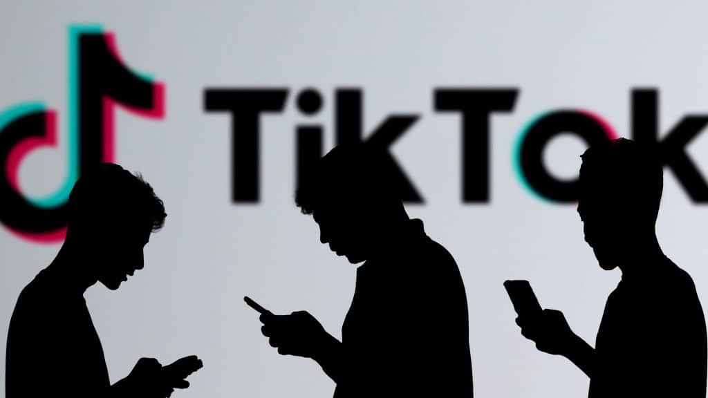 Silhueta de três pessoas usando celular; ao fundo, tela grande exibe letreiro do TikTok