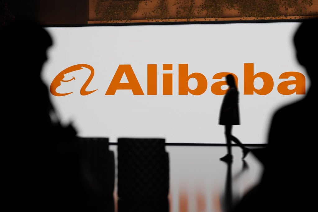 Logo da Alibaba ao fundo, com sombras de pessoas e outros objetos à frente