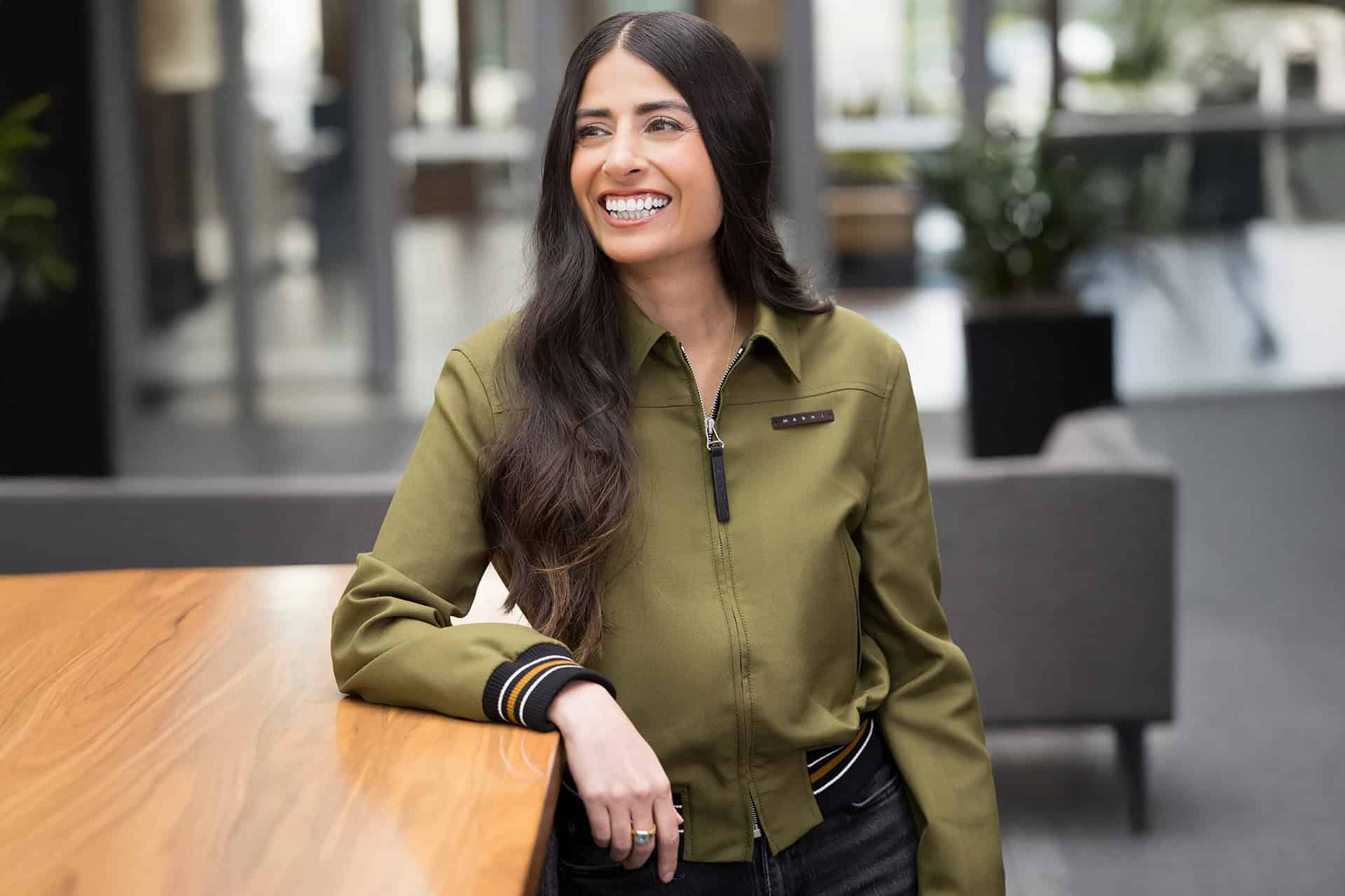 Asha Sharma sorrindo e com um braço apoiado em uma mesa
