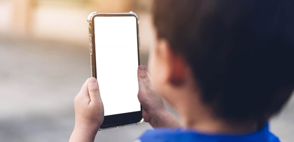 à frente, desfocado, o rosto de uma criança; à frente um smartphone que ela está segurando com a tela branca