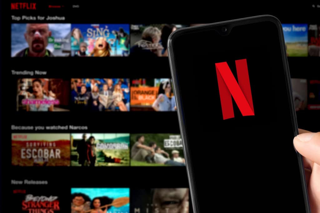 Ao fundo, desfocado, tela inicial da Netflix em uma TV; à frente, celular com o logo da empresa na tela