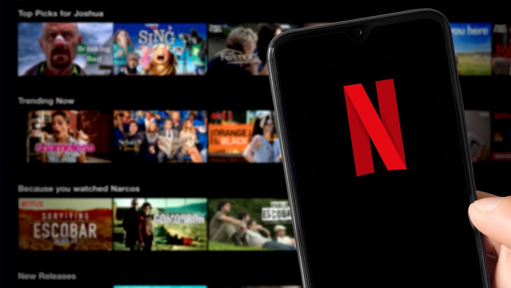 Ao fundo, desfocado, tela inicial da Netflix em uma TV; à frente, celular com o logo da empresa na tela