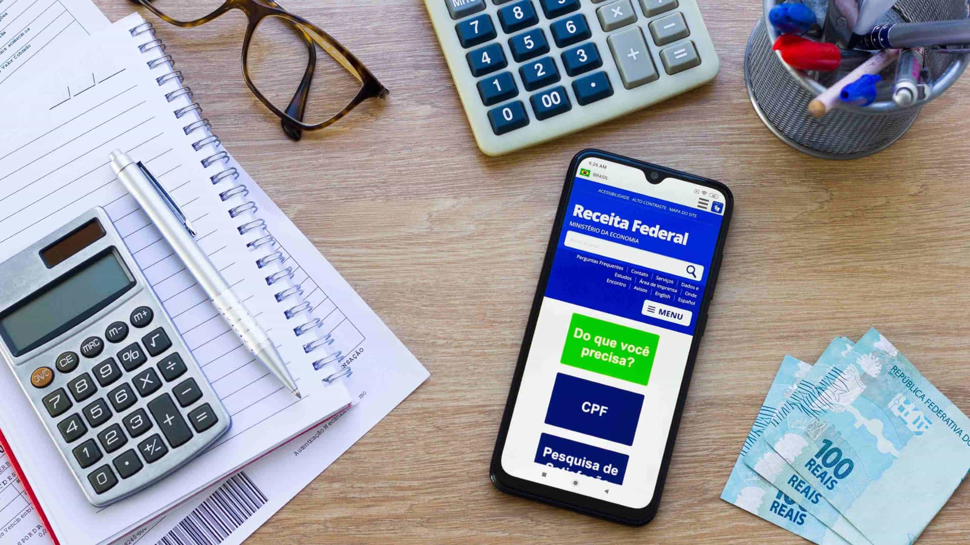 Mesa com calculadora e celular aberto no app da Receita Federal para calcular o imposto de renda (IR)