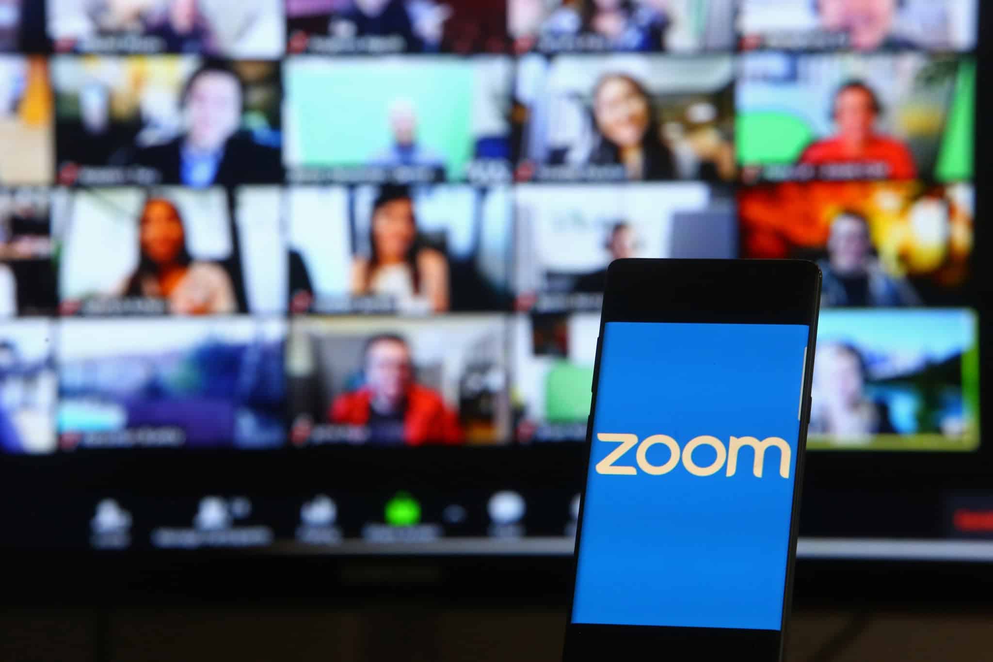 Ao fundo, borrado, a tela do Zoom com várias pessoas na chamda; à frente, logo da empresa em um smartphone